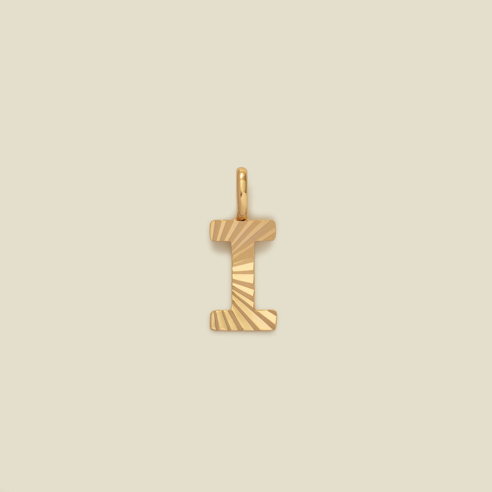 A small gold I-shaped charm with a textured, sunburst striped pattern, shown on a solid light beige background.