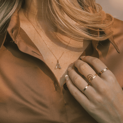 A person in a brown satin button-up top wearing a delicate gold pendant chain necklace with an initial 'A' charm and multiple stacked gold rings.