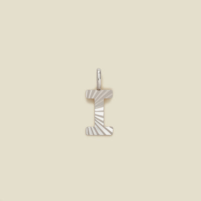 A small silver I-shaped charm with a textured, sunburst striped pattern, shown on a solid light beige background.