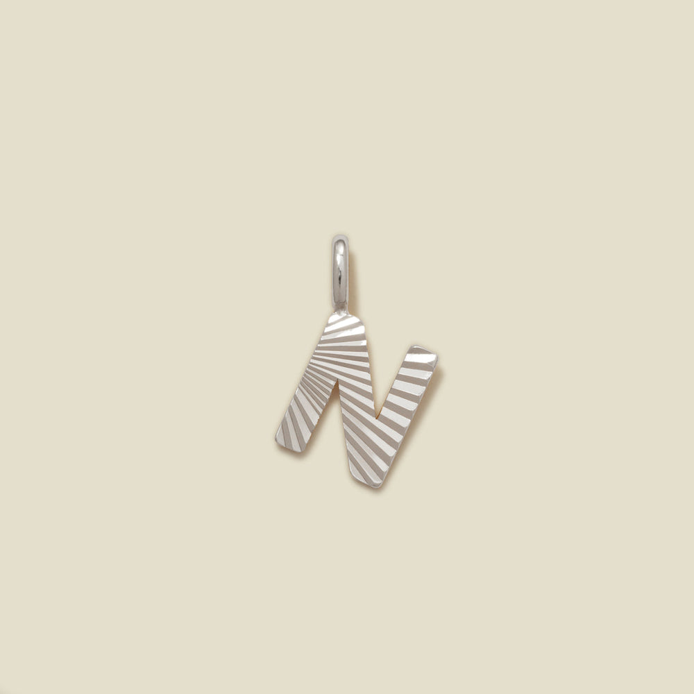 A small silver N-shaped charm with a textured, sunburst striped pattern, shown on a solid light beige background.