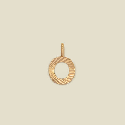 A small gold O-shaped charm with a textured, sunburst striped pattern, shown on a solid light beige background.