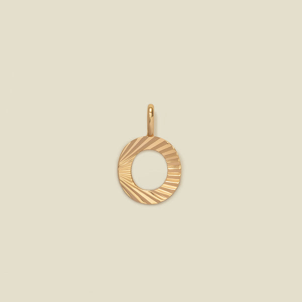 A small gold O-shaped charm with a textured, sunburst striped pattern, shown on a solid light beige background.