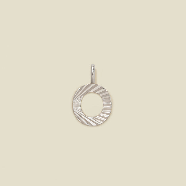 A small silver O-shaped charm with a textured, sunburst striped pattern, shown on a solid light beige background.