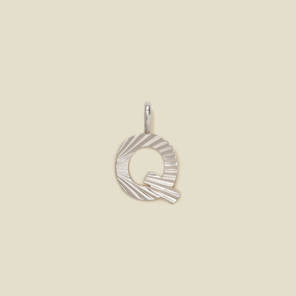A small silver Q-shaped charm with a textured, sunburst striped pattern, shown on a solid light beige background.