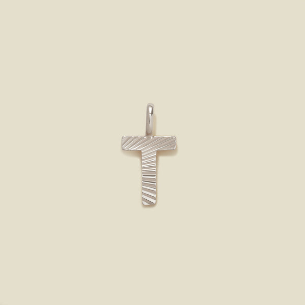 A small silver T-shaped charm with a textured, sunburst striped pattern, shown on a solid light beige background.