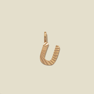 A small gold U-shaped charm with a textured, sunburst striped pattern, shown on a solid light beige background.