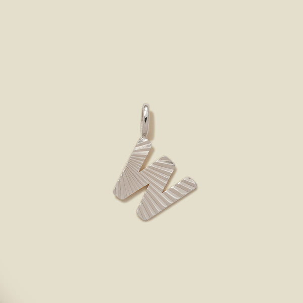 A small silver W-shaped charm with a textured, sunburst striped pattern, shown on a solid light beige background.
