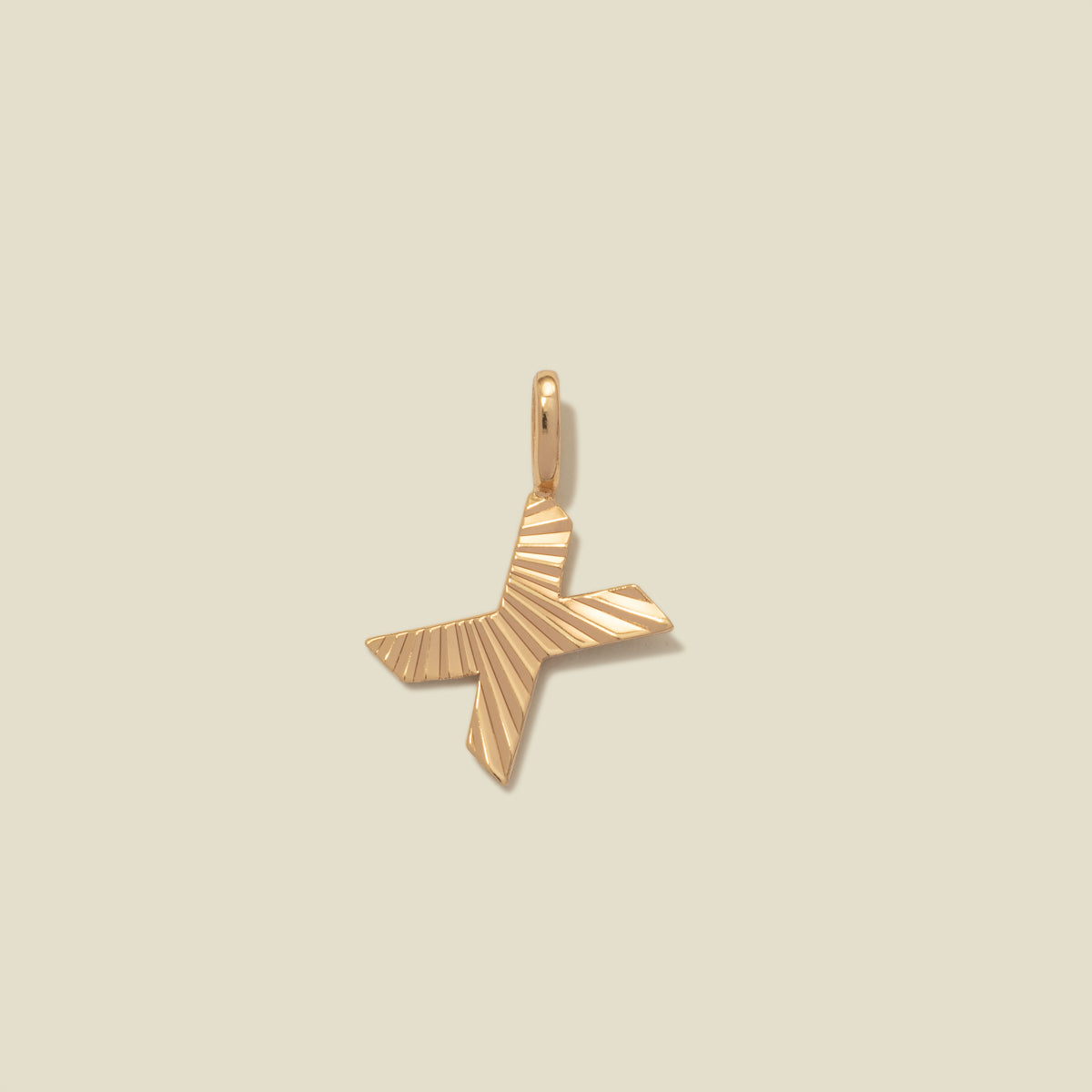 A small gold X-shaped charm with a textured, sunburst striped pattern, shown on a solid light beige background.