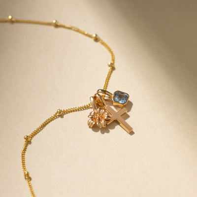 A dainty gold chain necklace with stationary round beads throughout, holding a gold flower charm, a gold cross charm, and a square light blue gemstone charm.