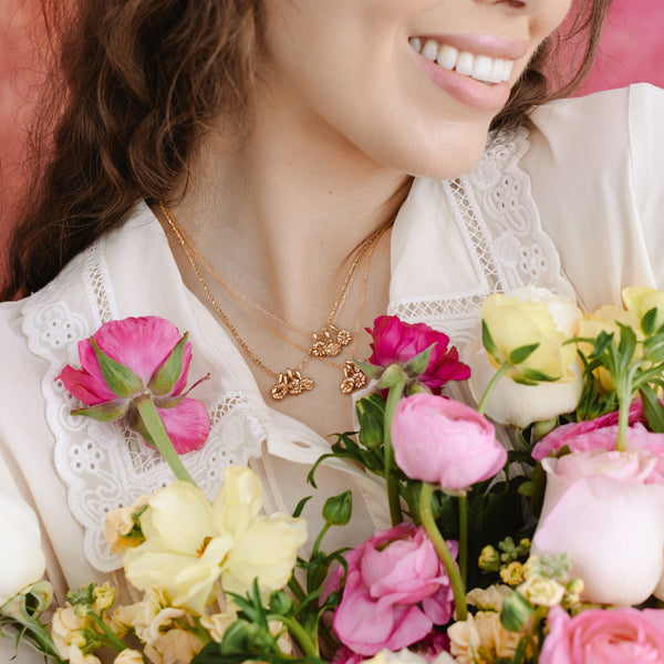 A close-up of a woman in a white linen top holding a bouquet of pink and yellow flowers, showcasing three separate dainty gold chain necklaces, each with multiple gold single flower charms.