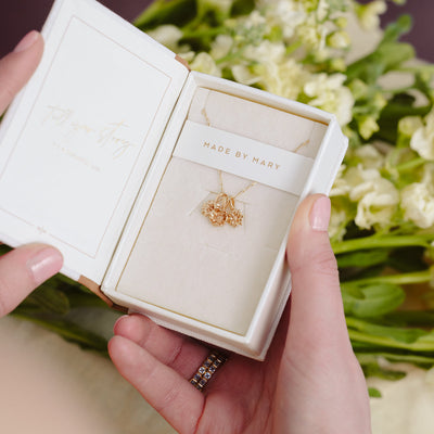 A person holding a small jewelry box with a delicate gold chain necklace inside, holding three different single flower charms. There is a necklace wrap across the top of the box with the words  'MADE BY MARY', and a bouquet of white flowers blurred in the background.