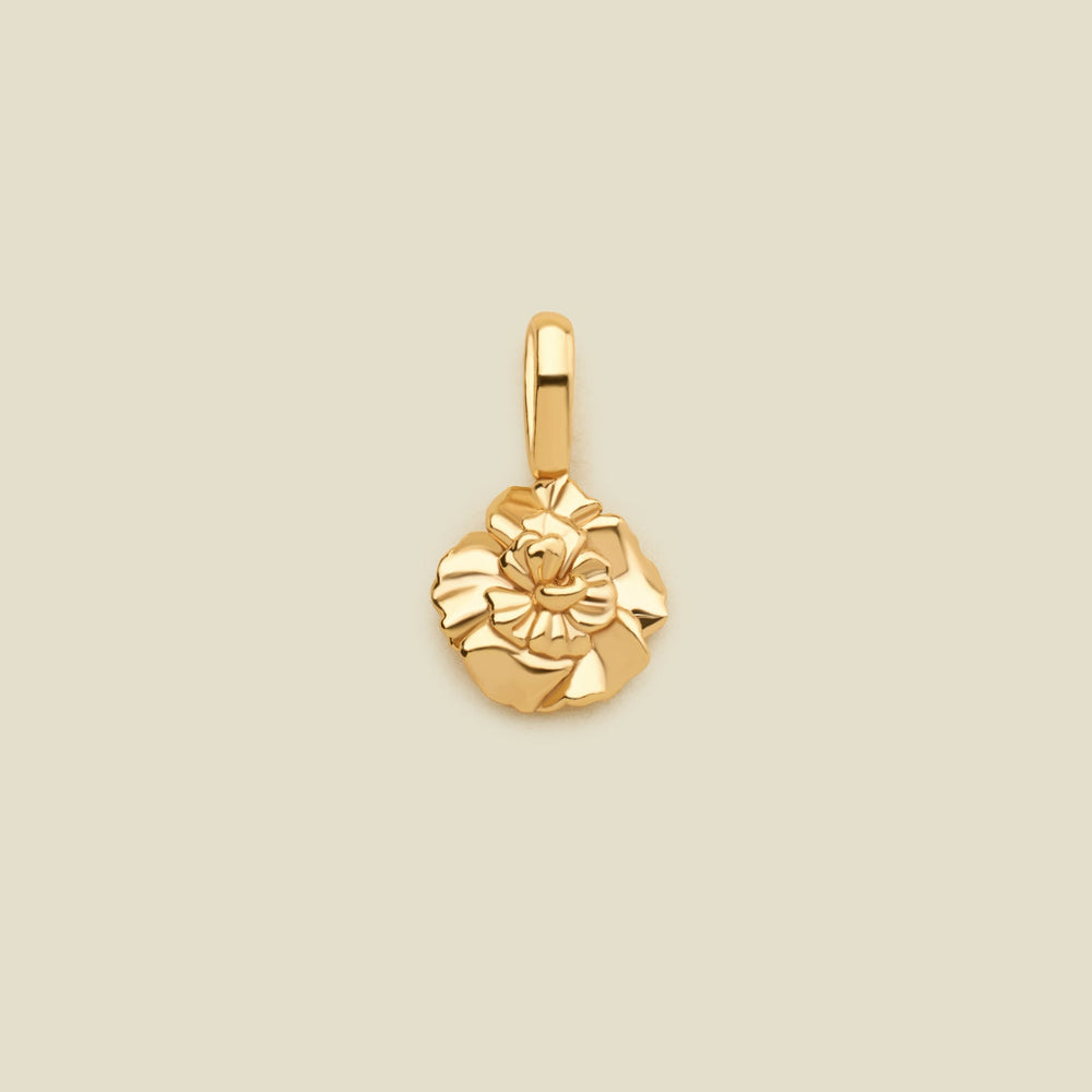 A polished yellow gold flower charm pendant with a textured, layered petal design and a top loop, centered on a solid light beige background.