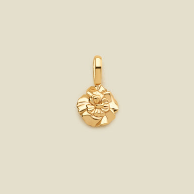 A polished yellow gold flower charm pendant with a textured, layered petal design and a top loop, centered on a solid light beige background.