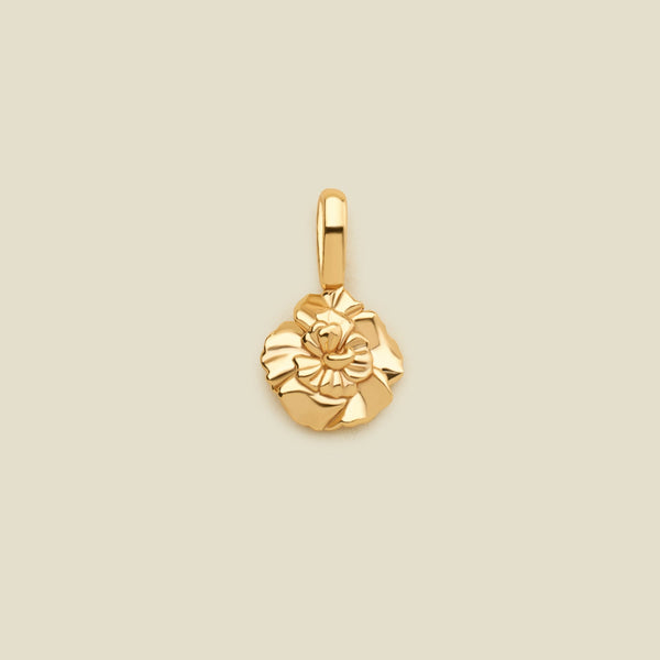 A polished yellow gold flower charm pendant with a textured, layered petal design and a top loop, centered on a solid light beige background.