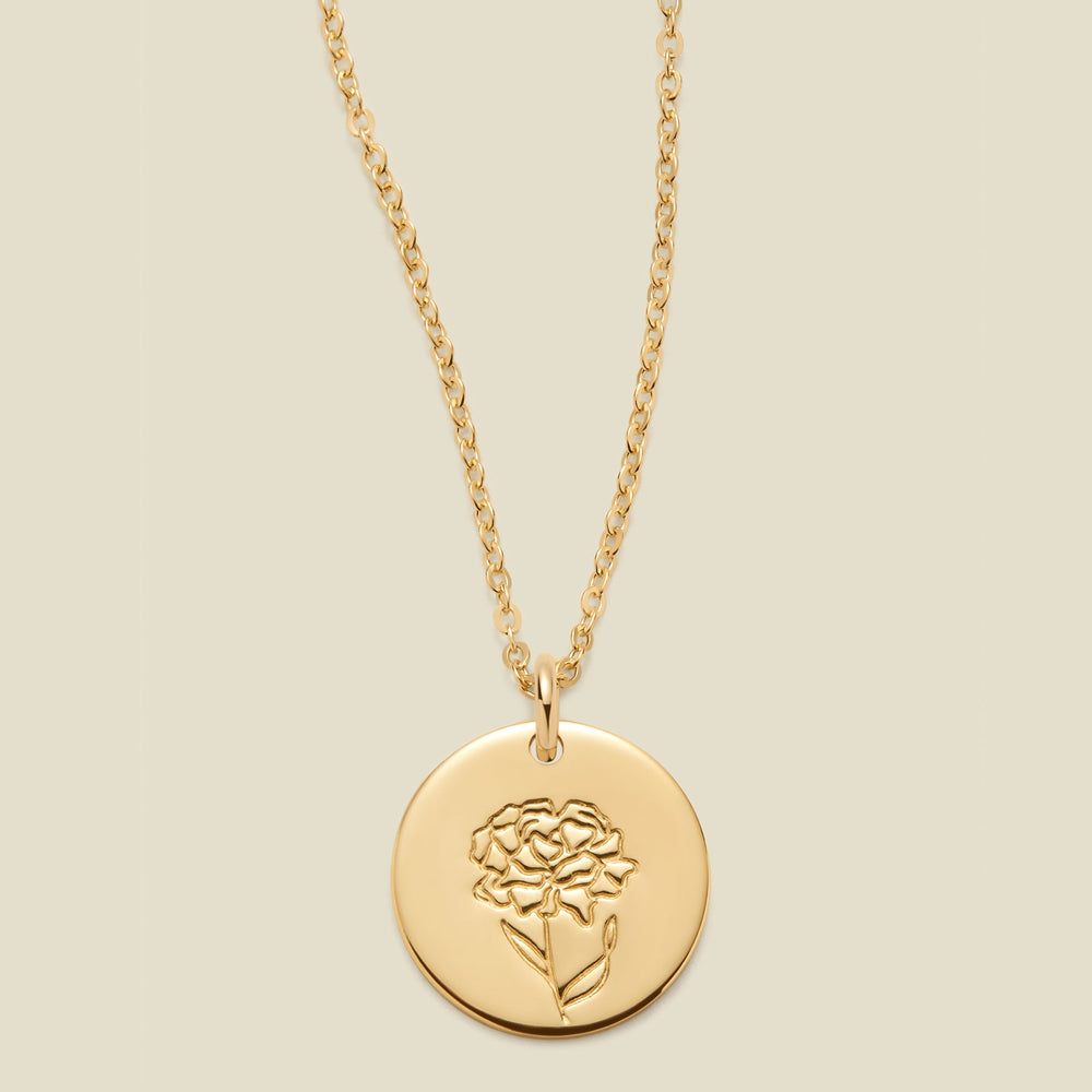 A gold chain necklace with a round gold disc pendant featuring an engraved carnation flower design, displayed against a solid light beige background.