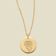 A gold chain necklace with a round gold disc pendant featuring an engraved carnation flower design, displayed against a solid light beige background.