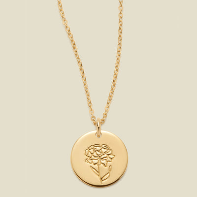A gold chain necklace with a round gold disc pendant featuring an engraved carnation flower design, displayed against a solid light beige background.