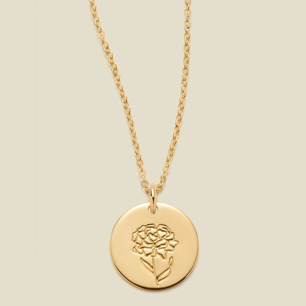 A gold chain necklace with a round gold disc pendant featuring an engraved carnation flower design, displayed against a solid light beige background.