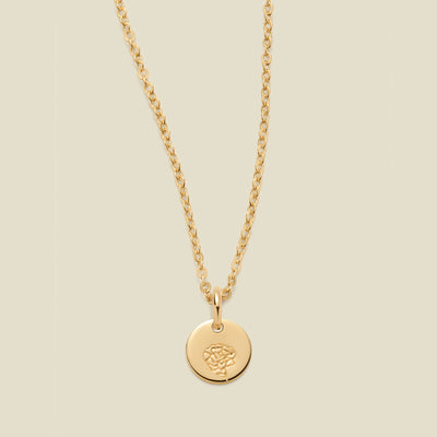 A gold chain necklace with a round gold disc pendant featuring an engraved carnation flower design, displayed against a solid light beige background.