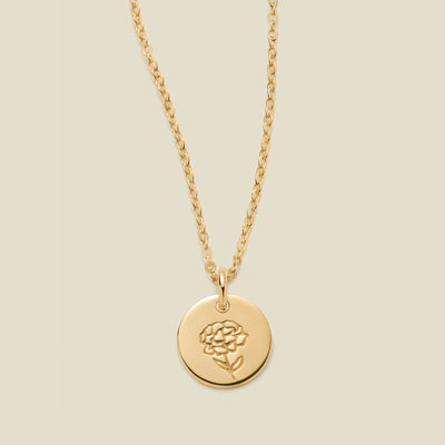 A gold chain necklace with a round gold disc pendant featuring an engraved carnation flower design, displayed against a solid light beige background.