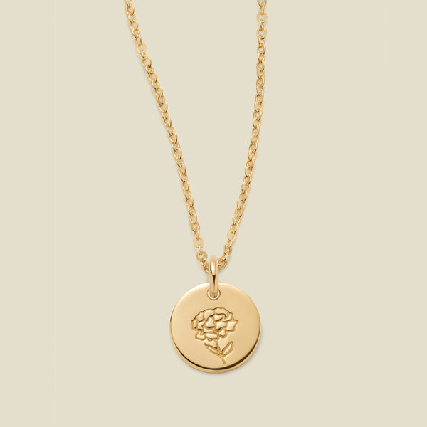 A gold chain necklace with a round gold disc pendant featuring an engraved carnation flower design, displayed against a solid light beige background.
