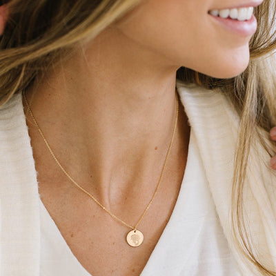 A close-up of a woman in a white top and cream cardigan wearing a delicate gold pendant chain necklace with a flat round disc. The disc is engraved with a carnation flower.