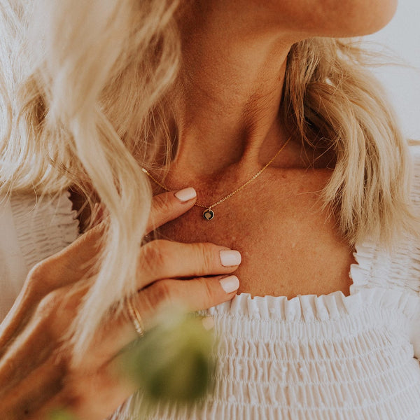 A close-up of a woman in a white pleated top wearing a delicate gold chain with a small round flat disc charm. The charm is engraved with a floral design.