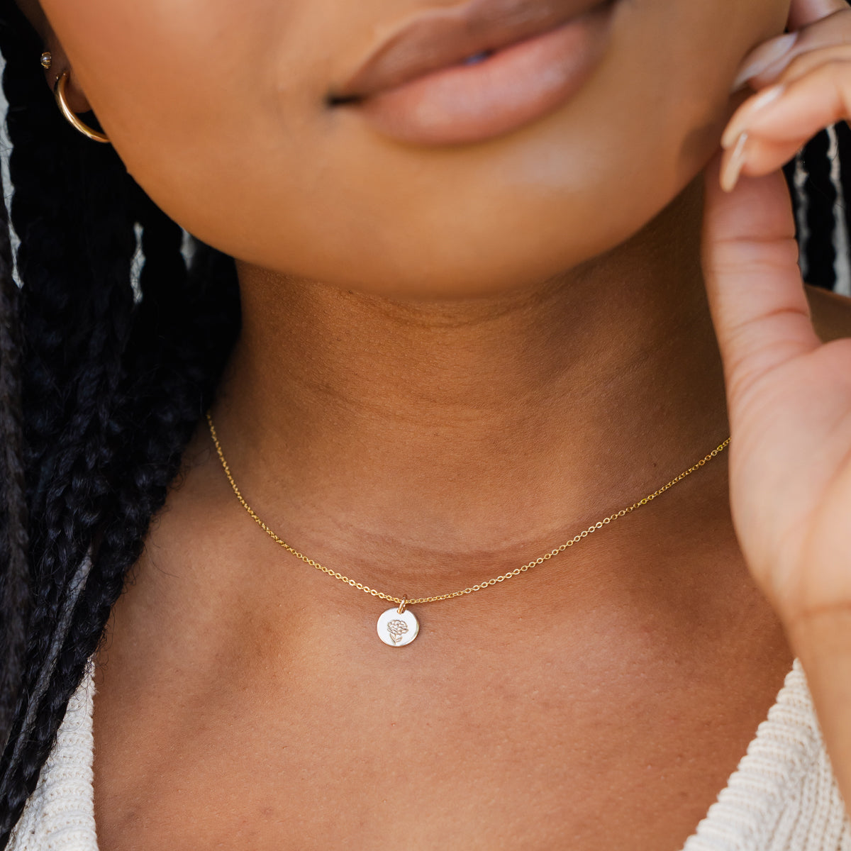 A close-up of a woman wearing a delicate gold pendant chain with a flat round disc. The disc is engraved with a carnation flower design.