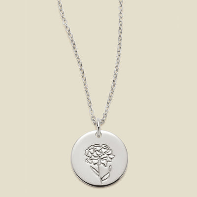 A silver chain necklace with a round silver disc pendant featuring an engraved carnation flower design, displayed against a solid light beige background.