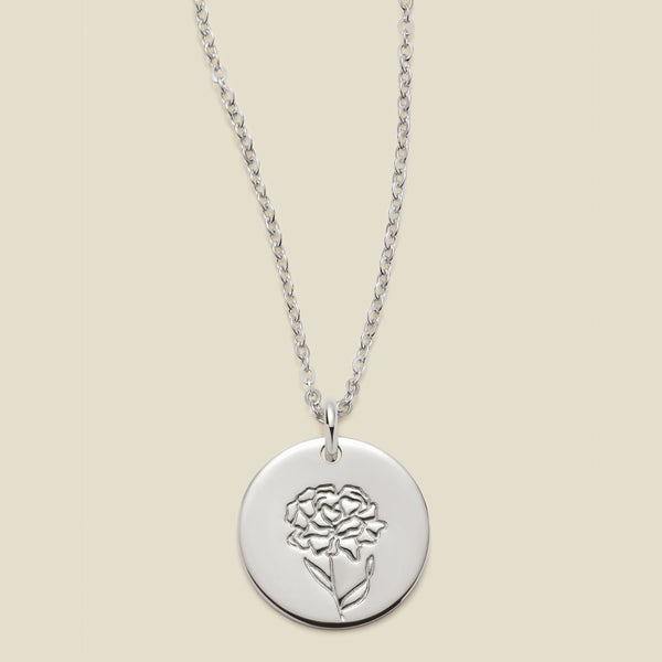 A silver chain necklace with a round silver disc pendant featuring an engraved carnation flower design, displayed against a solid light beige background.