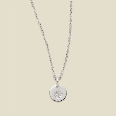 A silver chain necklace with a round silver disc pendant featuring an engraved carnation flower design, displayed against a solid light beige background.