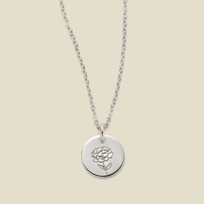 A silver chain necklace with a round silver disc pendant featuring an engraved carnation flower design, displayed against a solid light beige background.