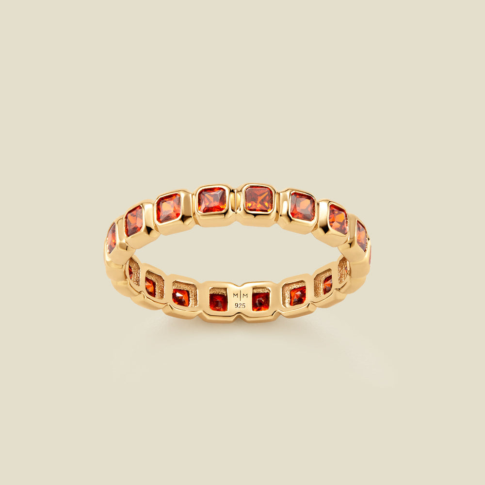 A gold eternity ring featuring a continuous row of square cut red-orange gemstones, shown on a solid light beige background.