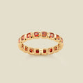 A gold eternity ring featuring a continuous row of square cut red-orange gemstones, shown on a solid light beige background.