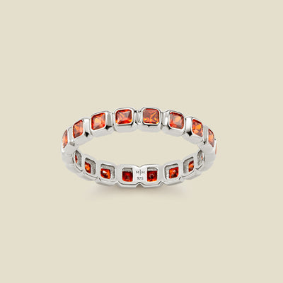 A silver eternity ring featuring a continuous row of square cut red-orange gemstones, shown on a solid light beige background.