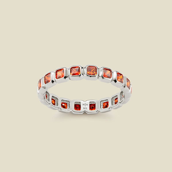 A silver eternity ring featuring a continuous row of square cut red-orange gemstones, shown on a solid light beige background.