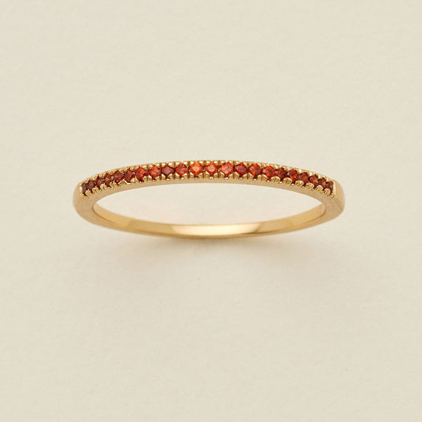 A gold ring featuring a single row of small round-cut red gemstones, photographed on a clean, solid, warm off-white background.
