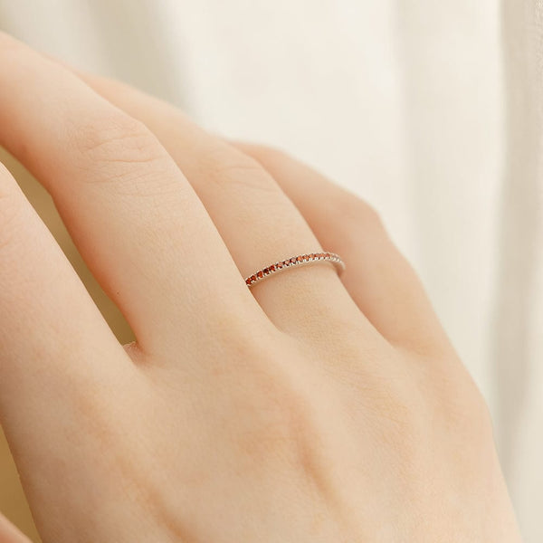 A close-up of a hand with a single thin silver ring, featuring a single row of red gemstones across the front, set against a bright white blurred background.