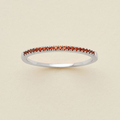A silver ring featuring a single row of small round-cut red gemstones, photographed on a clean, solid, warm off-white background.