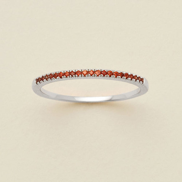A silver ring featuring a single row of small round-cut red gemstones, photographed on a clean, solid, warm off-white background.