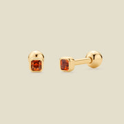 A pair of gold post earrings with square-cut red-orange gemstones, shown from front and side angles on a light beige background.