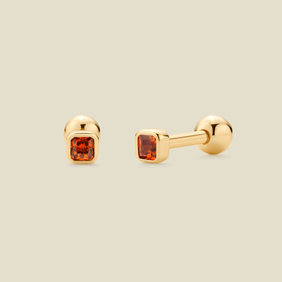 A pair of gold post earrings with square-cut red-orange gemstones, shown from front and side angles on a light beige background.