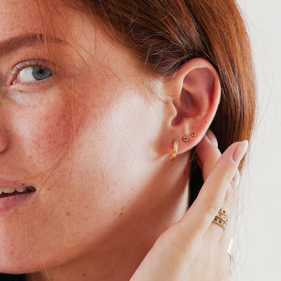 A close-up of a woman's ear wearing a polished gold hoop earring and two square gemstone studs, one a red-orange color and the other an amber orange.