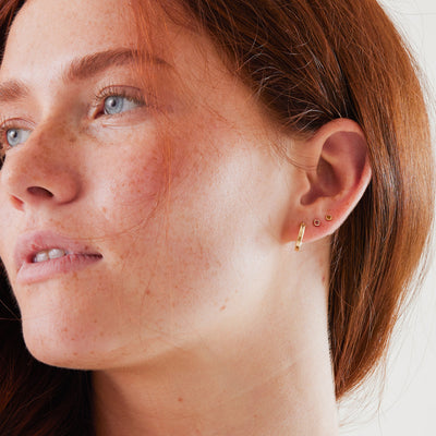 A close-up of a woman's ear wearing a polished gold hoop earring and two gold square gemstone stud earrings.