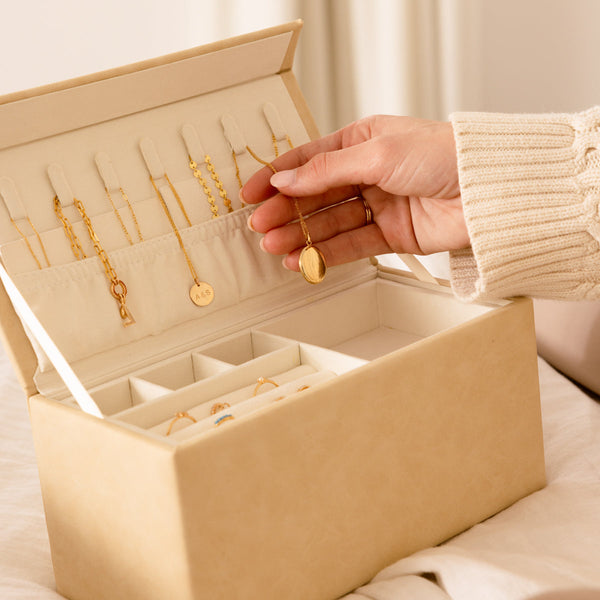 An open rectangular cream colored jewelry box, with multiple gold necklaces hanging from hooks inside the lid. A person is reaching in and holding out a gold locket necklace.