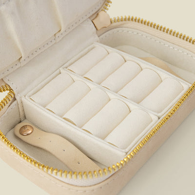 An open cream-colored jewelry organizer case with a gold zipper and white interior compartments, shown on a solid light beige background.