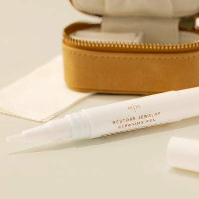A close-up of a white jewelry cleaner pean with the words 'RESTORE JEWELRY CLEANING PEN' in brown on the front, set in front of an open tan faux-leather jewelry case with gold-tone zipper and shown on a solid beige surface.