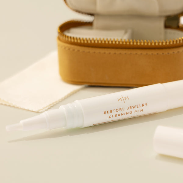 A close-up of a white jewelry cleaner pean with the words 'RESTORE JEWELRY CLEANING PEN' in brown on the front, set in front of an open tan faux-leather jewelry case with gold-tone zipper and shown on a solid beige surface.