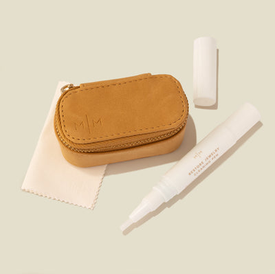A tan faux-leather jewelry case with gold-tone zipper, shown with a white polishing cloth and a white jewelry cleaner applicator pen on a soft beige background.