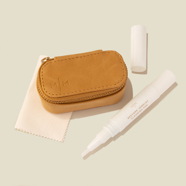 A tan faux-leather jewelry case with gold-tone zipper, shown with a white polishing cloth and a white jewelry cleaner applicator pen on a soft beige background.